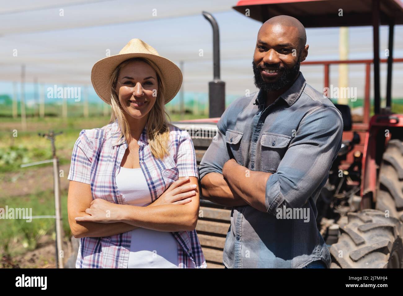 Lächelnder afroamerikanischer Mann und kaukasische Frau mit gekreuzten Armen, die vom Traktor im Gewächshaus stehen Stockfoto