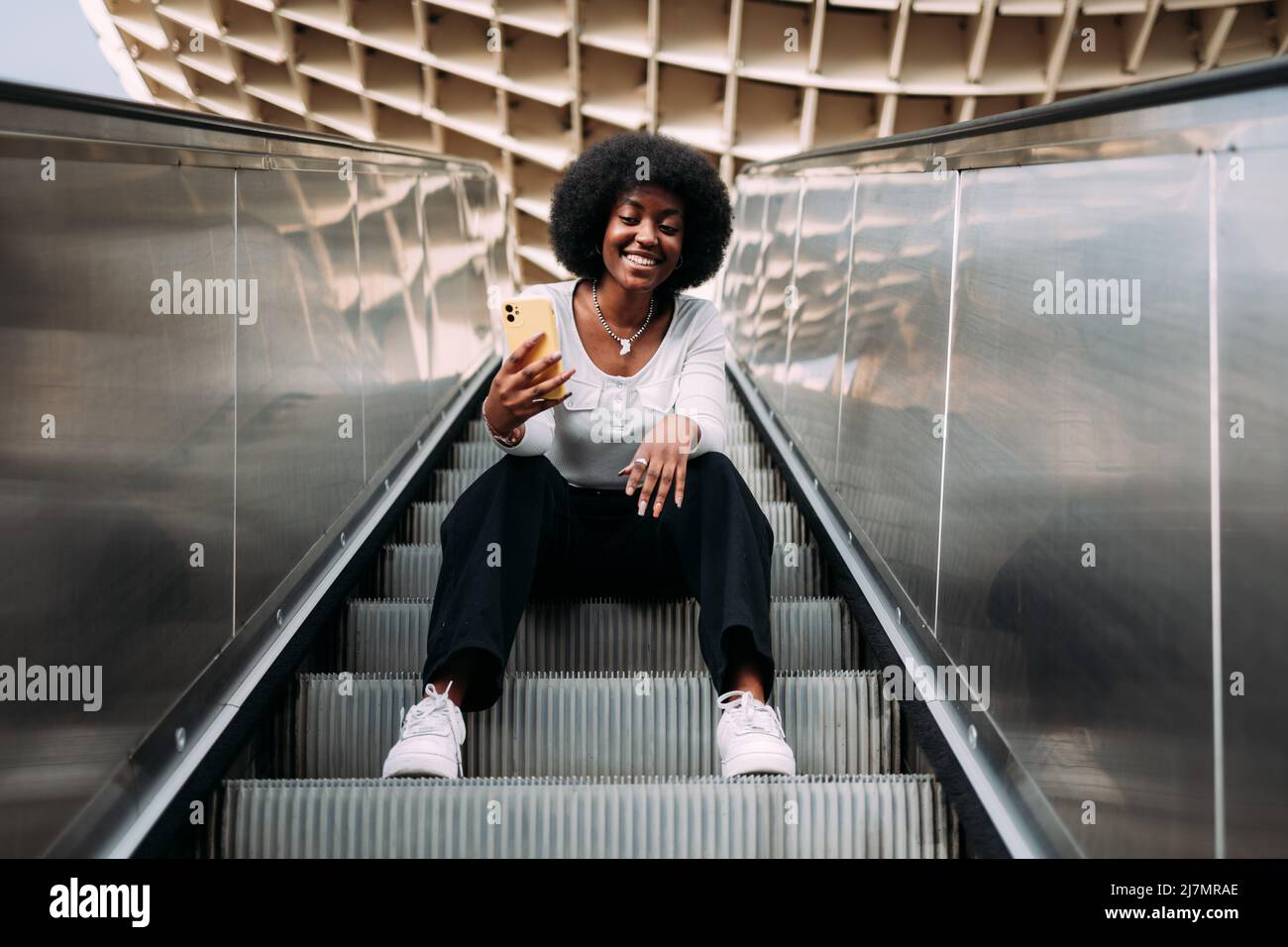 Lächelnde junge schwarze Teenagerin mit Afro-Haaren sitzt auf einer Rolltreppe im Freien und nimmt ein Selfie auf. Stockfoto