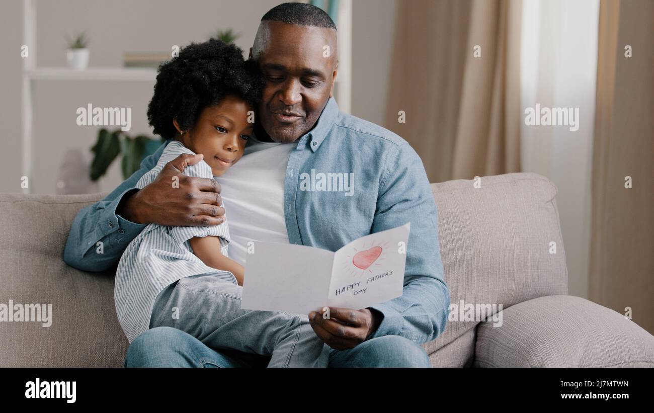 Reifer afroamerikanischer Mann mit niedlichen Mädchen umarmt sitzen im Zimmer auf Sofa Vater liest Grußkarte liebevolle Tochter gemacht Geschenk mit eigenen Händen für Stockfoto