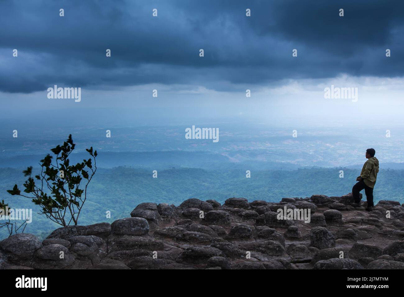 Ein Mann in einer Regenjacke steht am Rand einer Klippe und blickt auf die dunklen Regenwolken über den Bergen, Felsformationen, die von der Natur aufgetreten sind. Stockfoto