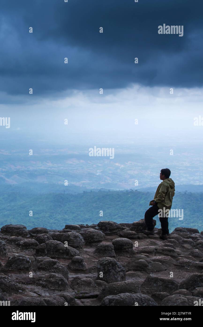 Ein Mann in einer Regenjacke steht am Rand einer Klippe und blickt auf die dunklen Regenwolken über den Bergen, Felsformationen, die von der Natur aufgetreten sind. Stockfoto