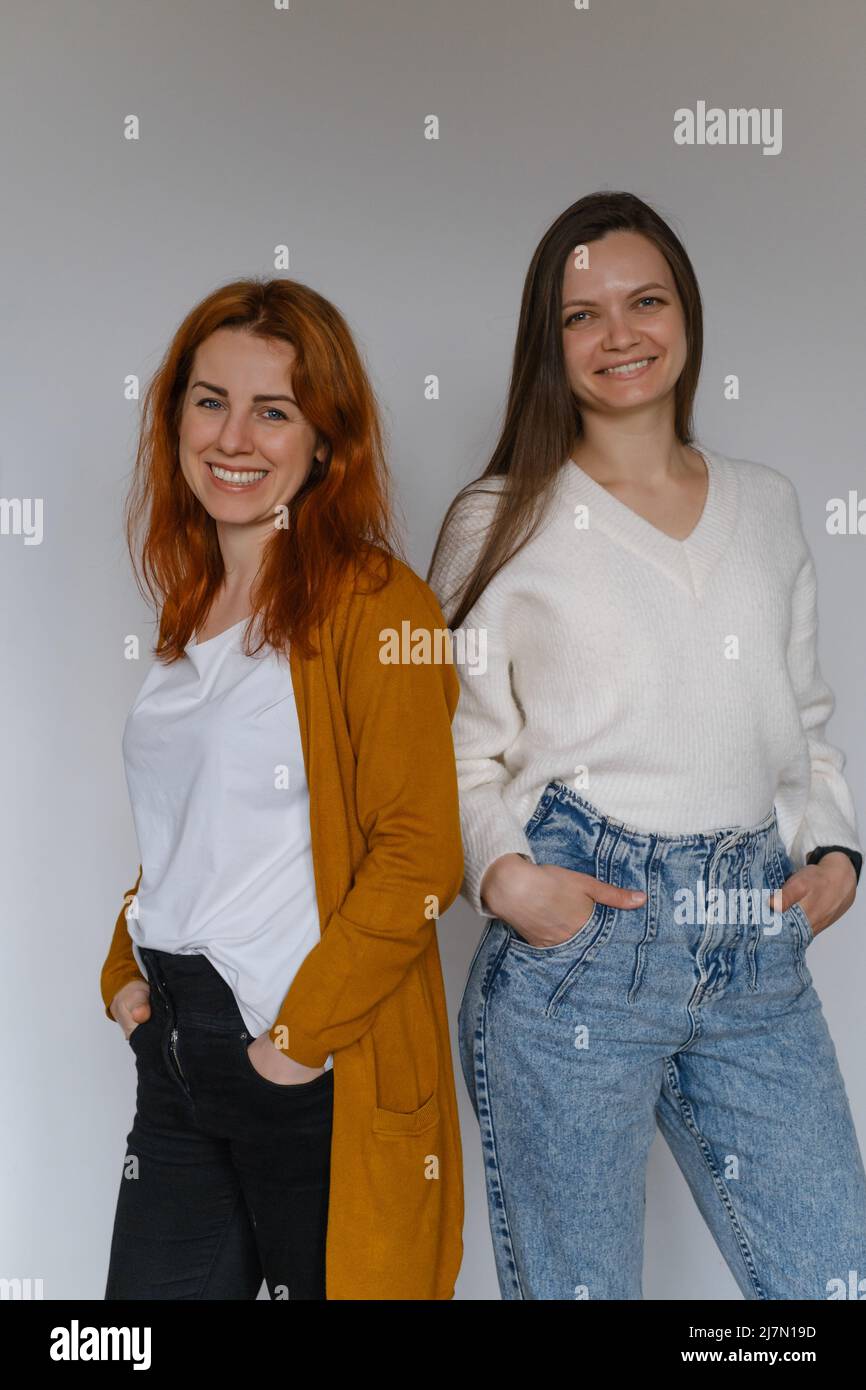 Glückliche zwei Frauen im Alter von 20s 40s über einer weißen Wand. Mitarbeiter in legerer Kleidung, die auf die Kamera schauen. Frau mit langen Haaren, fröhliche Freiberuflerin Stockfoto