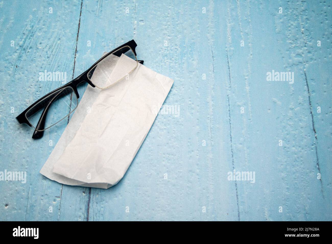 Draufsicht auf Lesebrille mit schwarzen Bügeln und Brillenreinigungstuch auf hellblau lackierter Holzfläche Stockfoto