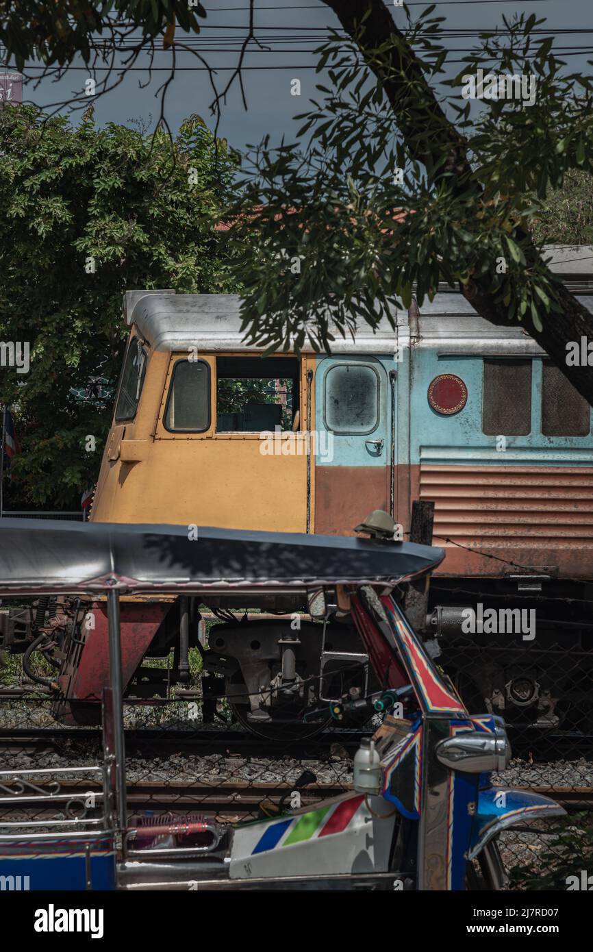 Bangkok, Thailand - 29. Apr 2022 : Seitenansicht des Blue Tuk Tuk (3-Rad-Taxi) und der Diesel-E-Lokomotive, die überlappend auf einer alten Straße abgestellt wird. Wählen Sie Stockfoto