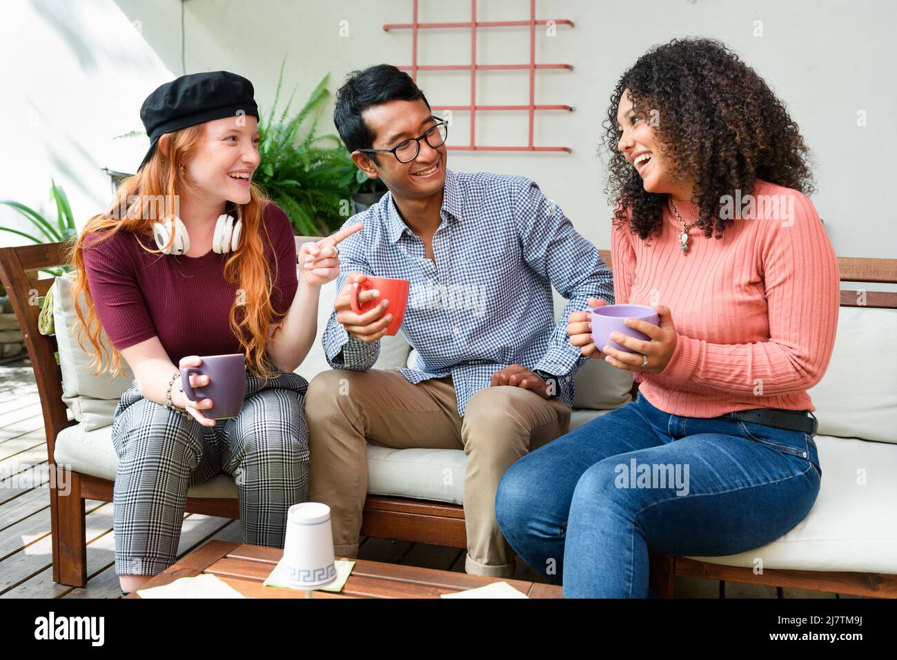 Eine Gruppe von multirassischen Freunden mit Tassen Kaffee plaudert und schaut sich gegenseitig an, während sie auf einem bequemen Sofa auf der hellen Terrasse sitzen Stockfoto