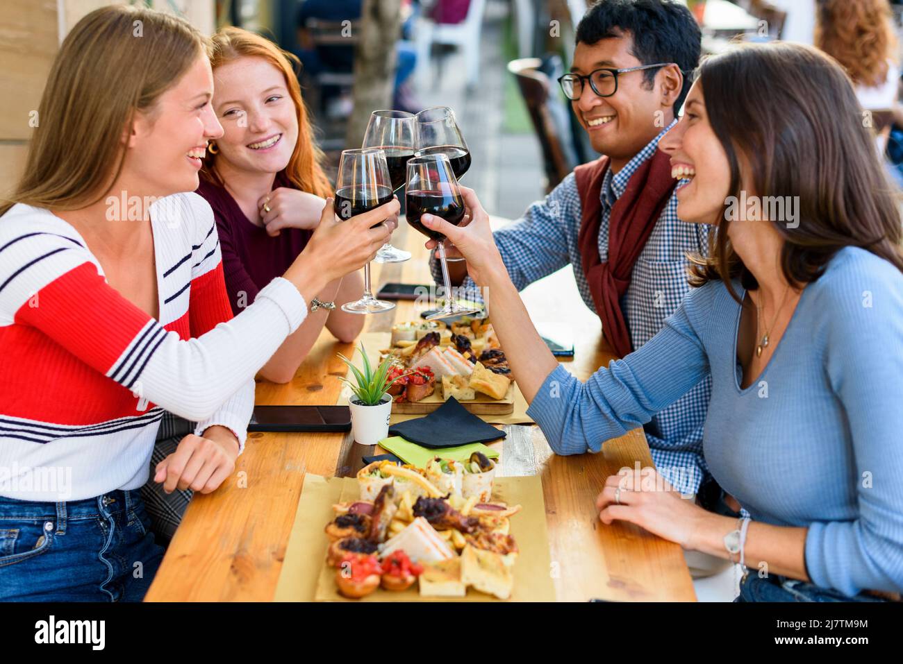 Eine Gruppe fröhlicher multirassischer Freunde, die sich gegenseitig angucken und mit Wein in die Gläser klirren, während sie ein festliches Ereignis am Tisch mit Essen auf der Terra feiern Stockfoto