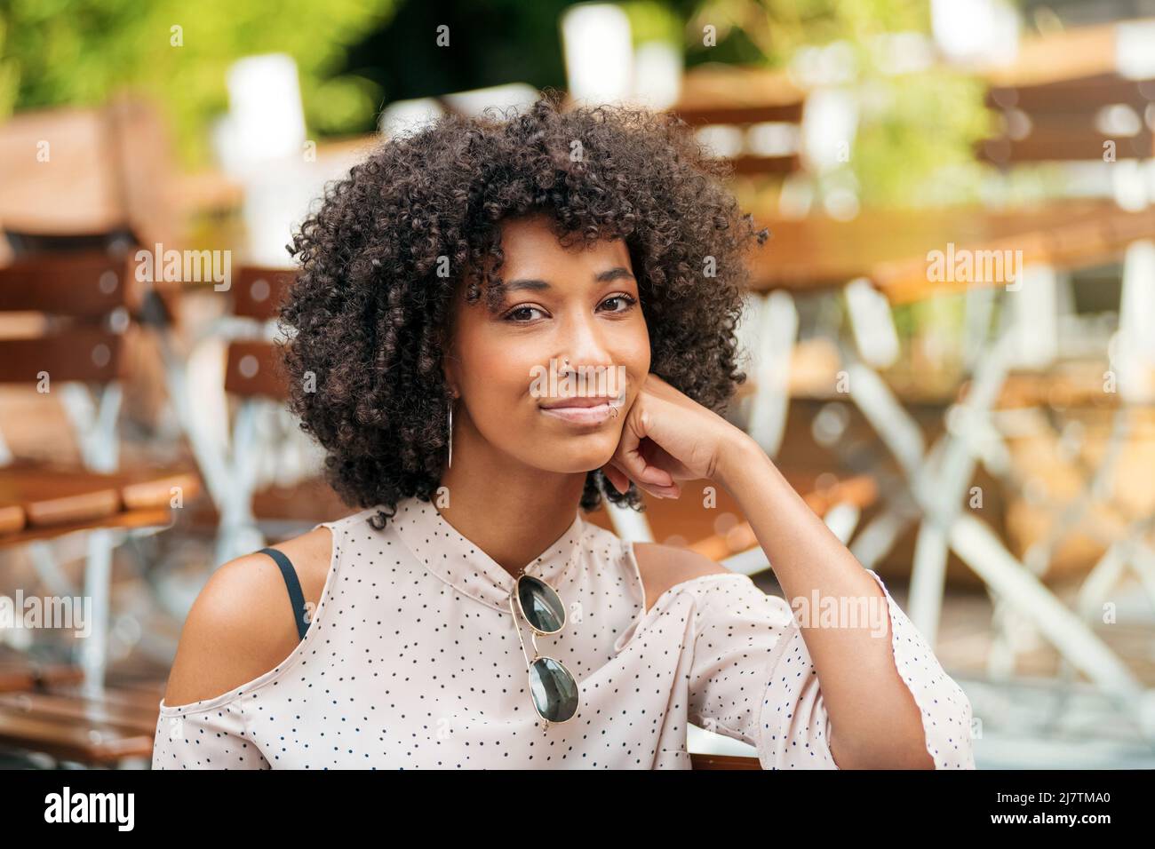 Attraktive afroamerikanische Frau mit lockigen Haaren, die auf die Kamera schaut, während sie auf der Terrasse des Cafés mit Tischen auf verschwommenem Hintergrund sitzt Stockfoto