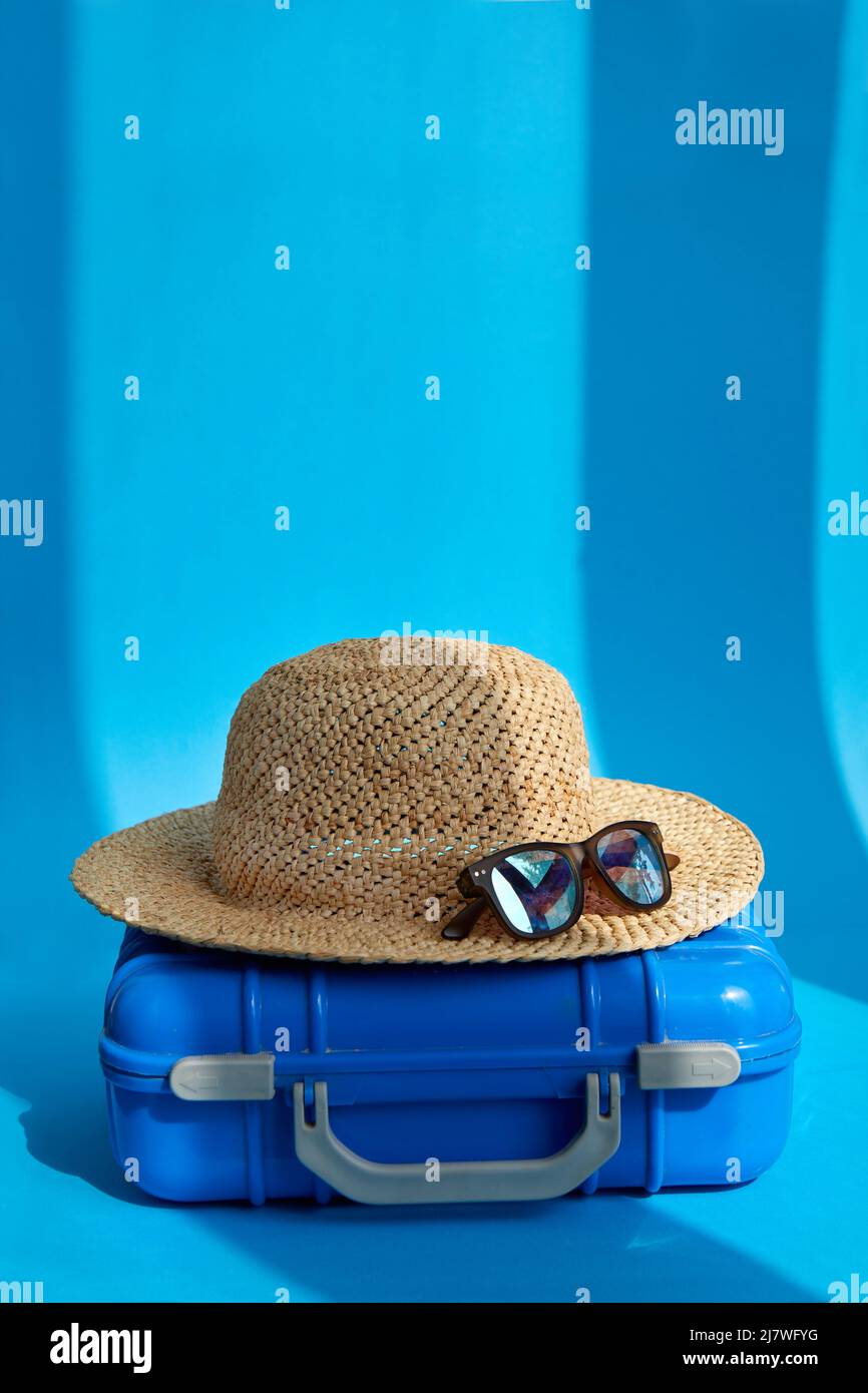 Blauer Plastikkoffer mit Strohhut und Sonnenbrille auf dem Boden in einem hellen Studio für den Sommerurlaub platziert Stockfoto