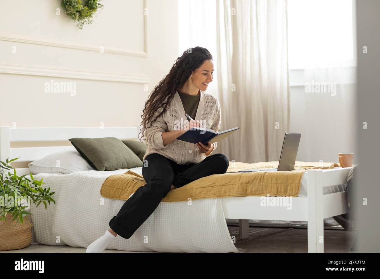 Lächelnde Frau sitzt auf dem Bett und schreibt mit dem pc im Notebook Stockfoto