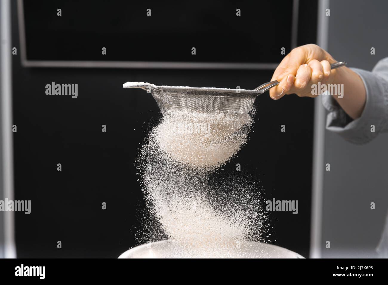 Nahaufnahme einer menschlichen Hand, die Mehl sieben und sich auf dunklem Hintergrund zum Backen vorbereiten. Stockfoto