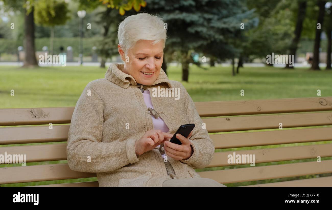 Überglücklich grauhaarige reife Frau, die im Freien sitzt, Smartphone-Apps nutzt, den Bildschirm anschaut, ältere Frau, die mit Verwandten chattet Stockfoto