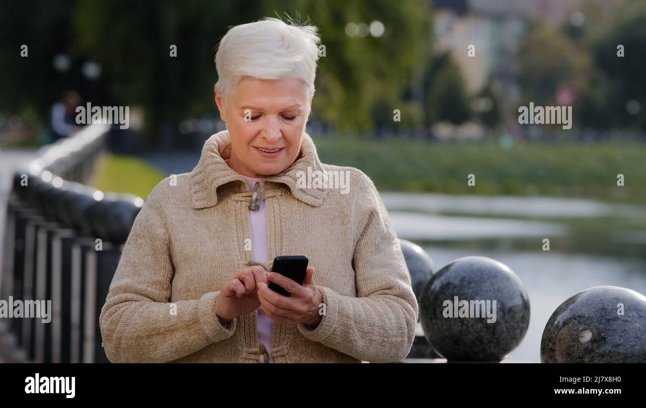 Lächelnd reifen 60s Frau SMS-Nachrichten auf moderne Smartphone-Gadget, glücklich Senior 70s Großmutter ruhen im Freien im Internet auf Handy surfen Stockfoto