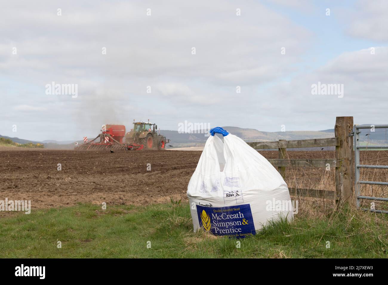 Ein Sack Saatgut am Eingang zu einem gepflügten Feld mit einem Traktor und einer Sämaschine im Hintergrund Stockfoto
