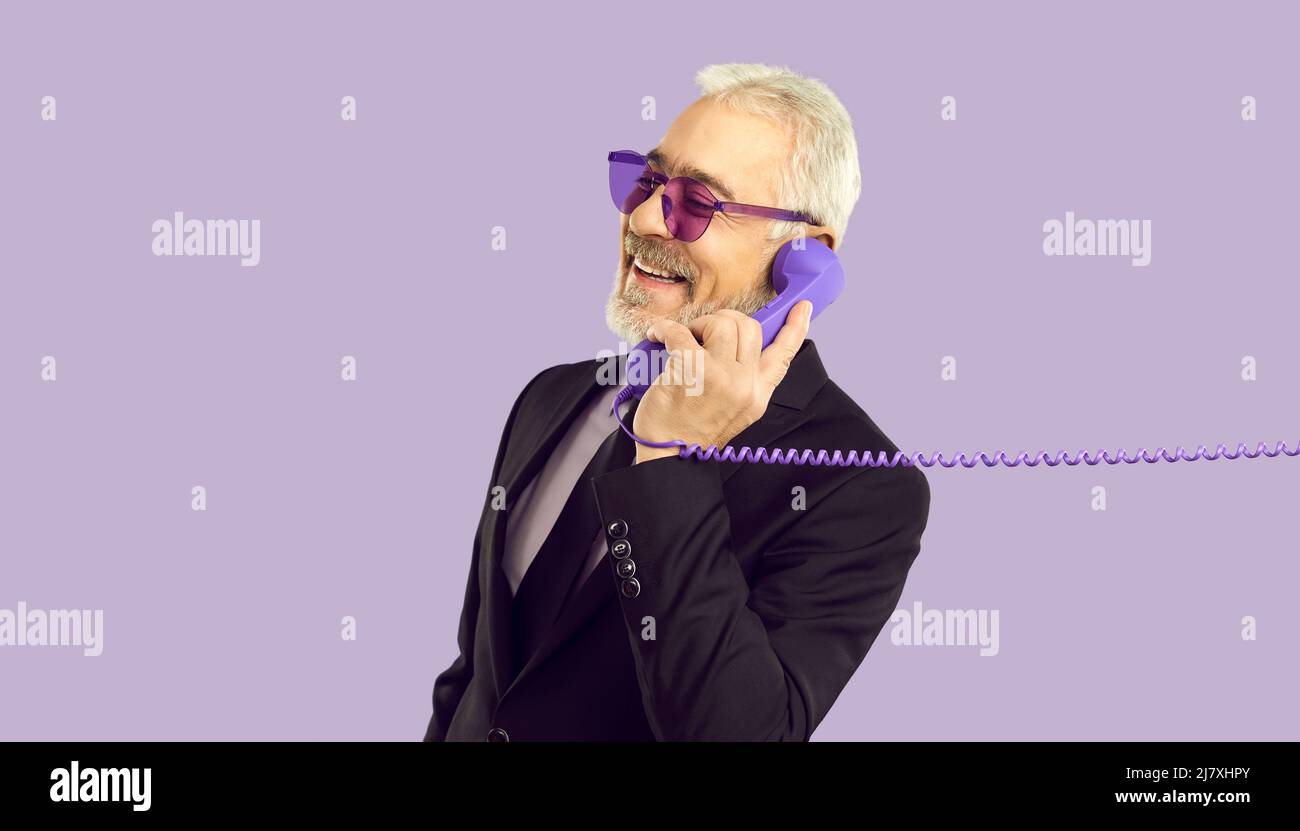 Fröhlicher, stilvoller und charismatischer älterer Mann, der auf einem Festnetztelefon auf purpurem Hintergrund spricht. Stockfoto