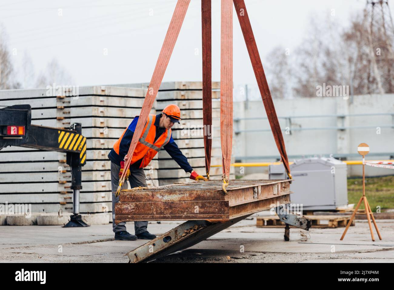 Slinger in Helm und Weste steuert das Entladen von Metallkonstruktionen auf der Baustelle. Weißer Handwerker entlädt Ladung. Stockfoto