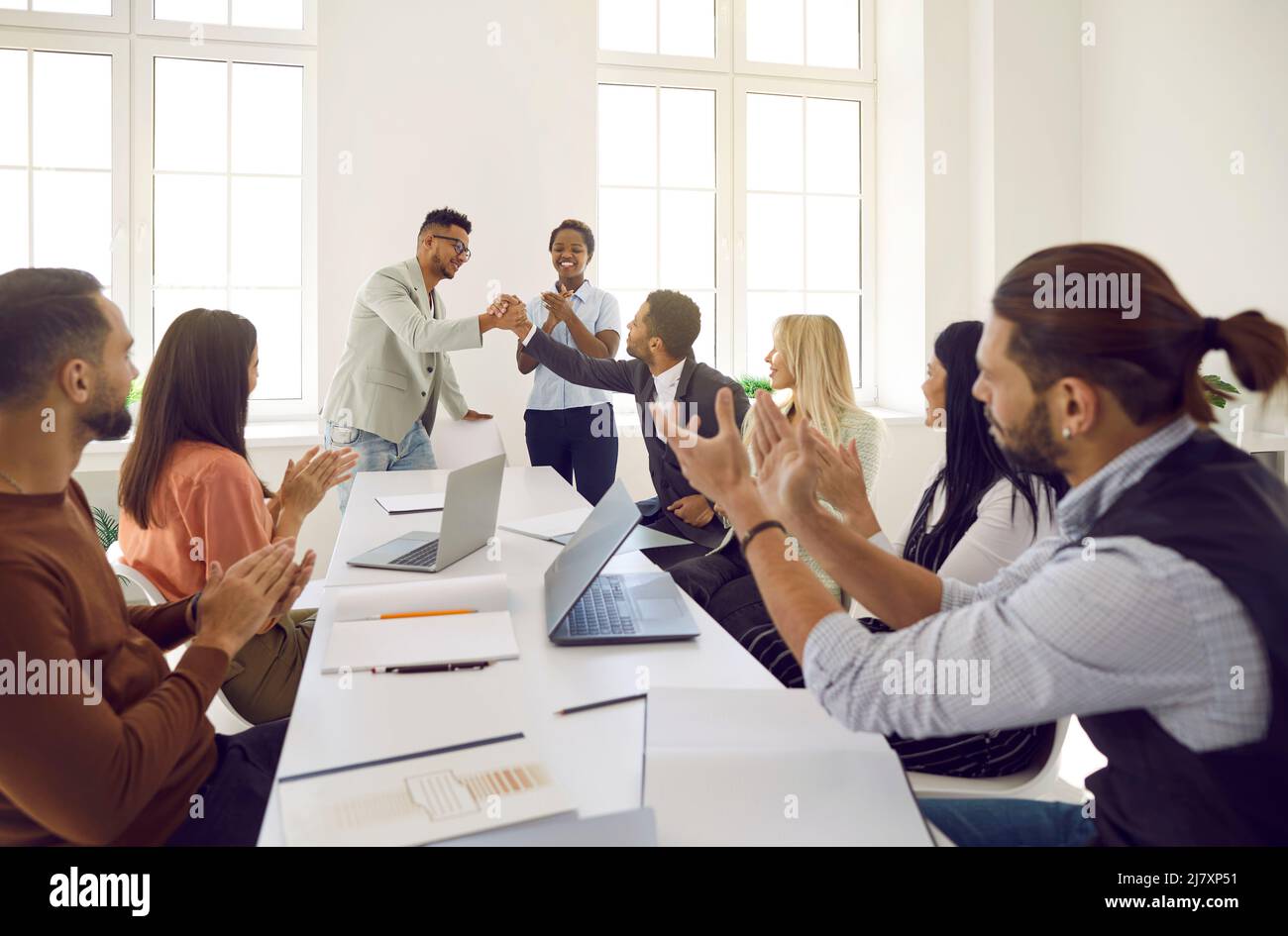 Ein glücklicher Mann schüttelt seinem Kollegen die Hände, während das vielfältige Geschäftsteam applaudiert Stockfoto