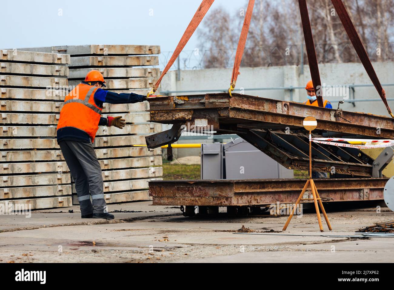 Slinger in Helm und Weste steuert das Entladen von Metallkonstruktionen auf der Baustelle. Weißer Handwerker entlädt Ladung. Stockfoto