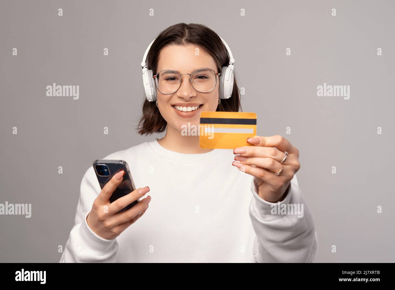 Lächelnde Frau hält eine Kreditkarte und ein Telefon in der Hand, während sie Kopfhörer trägt. Studio auf grauem Hintergrund. Stockfoto