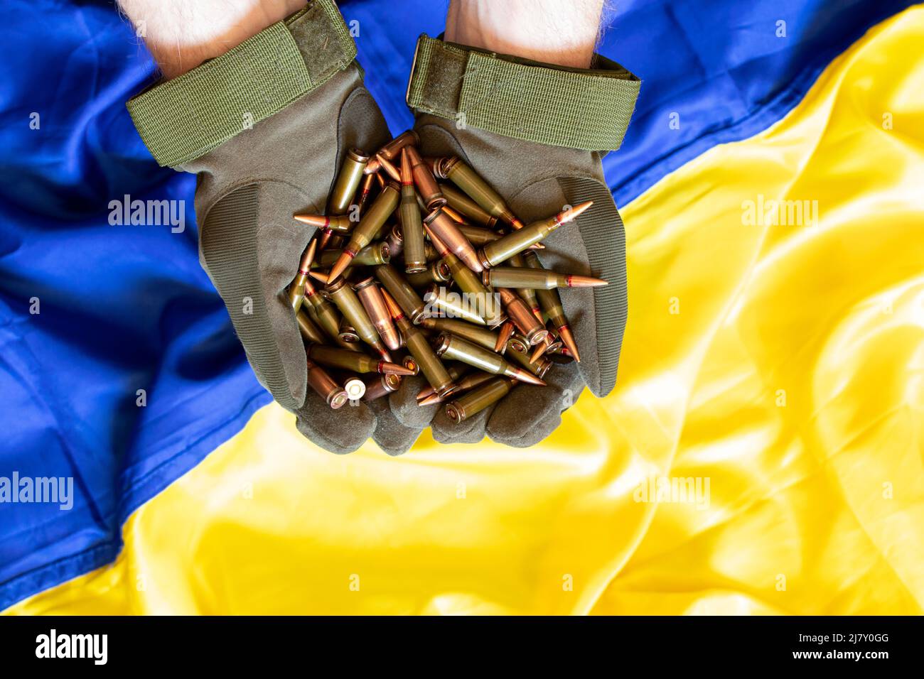 Ein ukrainischer Soldat in Handschuhen hält Kugeln aus einem Maschinengewehr vor dem Hintergrund der Flagge der Ukraine, Waffen der ukrainischen Armee, der Krieg i Stockfoto