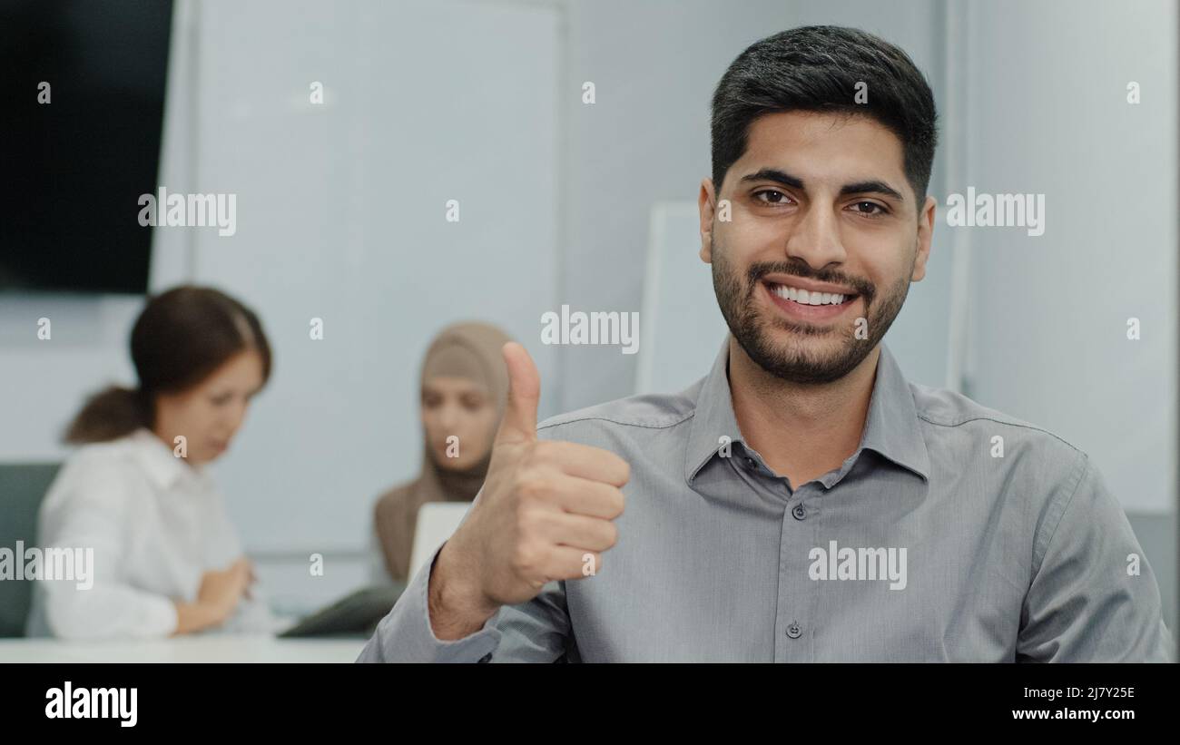 Porträt eines glücklich lächelnden arabischen männlichen Geschäftsführers, der den Daumen wie eine Geste vor der Kamera hochmacht. Der Mitarbeiter ist mit seiner Arbeit zufrieden und arbeitet mit einem großartigen Team zusammen Stockfoto