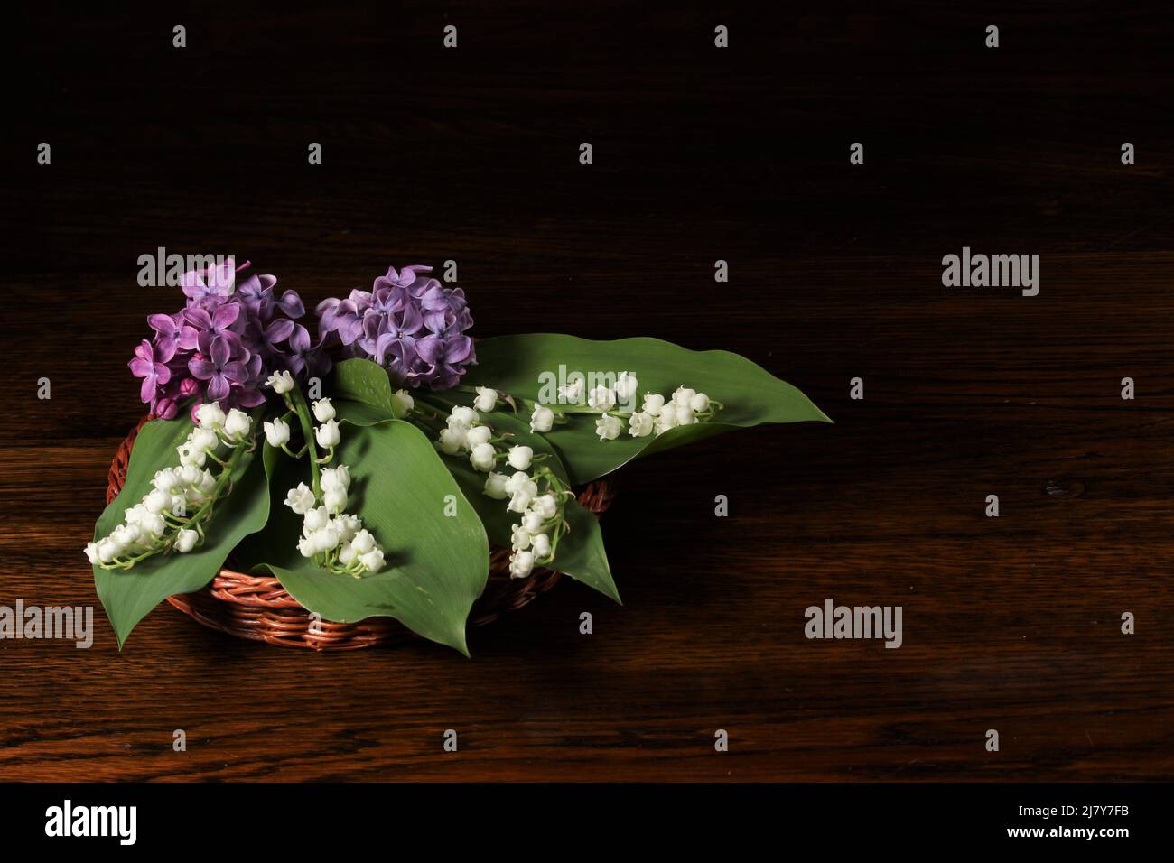 Schönes Bouquet von Maiglöckchen und lila Flieder in braunem Weidenkorb auf schwarzem Hintergrund. Visitenkarte. Stockfoto