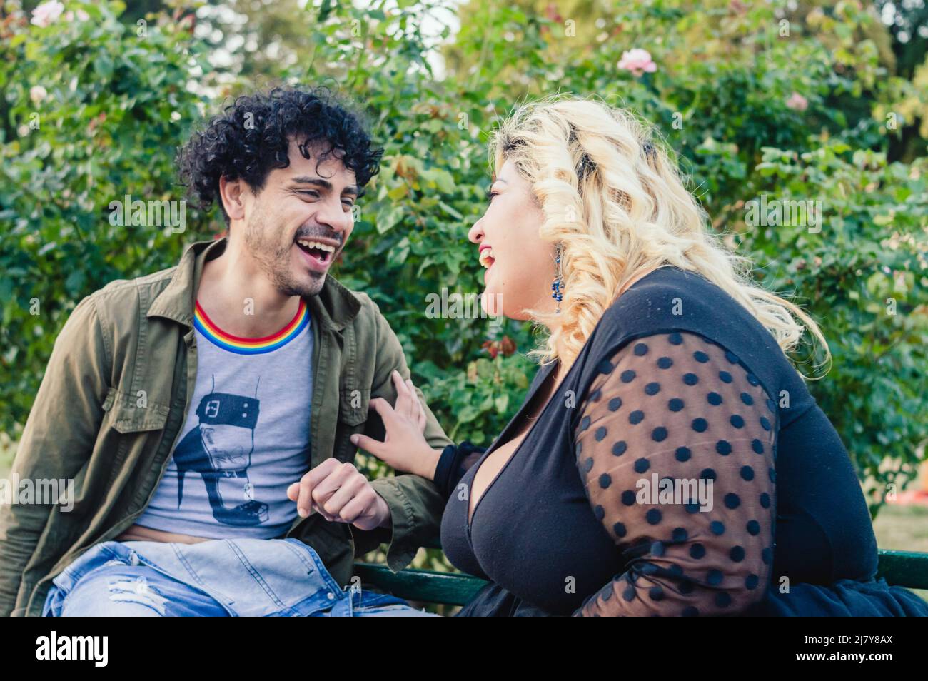 Mann mit Lockenwicklern und blonder Frau, junge kaukasische Freunde, sitzen im Freien, reden lachend und genießen zusammen. Stockfoto