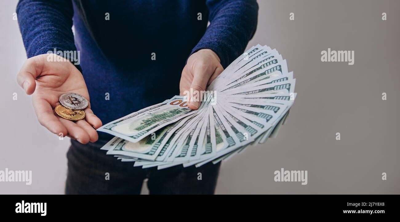 Ein Mann hält einen Fan von Hundert-Dollar-Scheinen und zwei Bitcoins. Austausch von Dollar gegen Kryptowährung, Kauf von Bitcoins Stockfoto