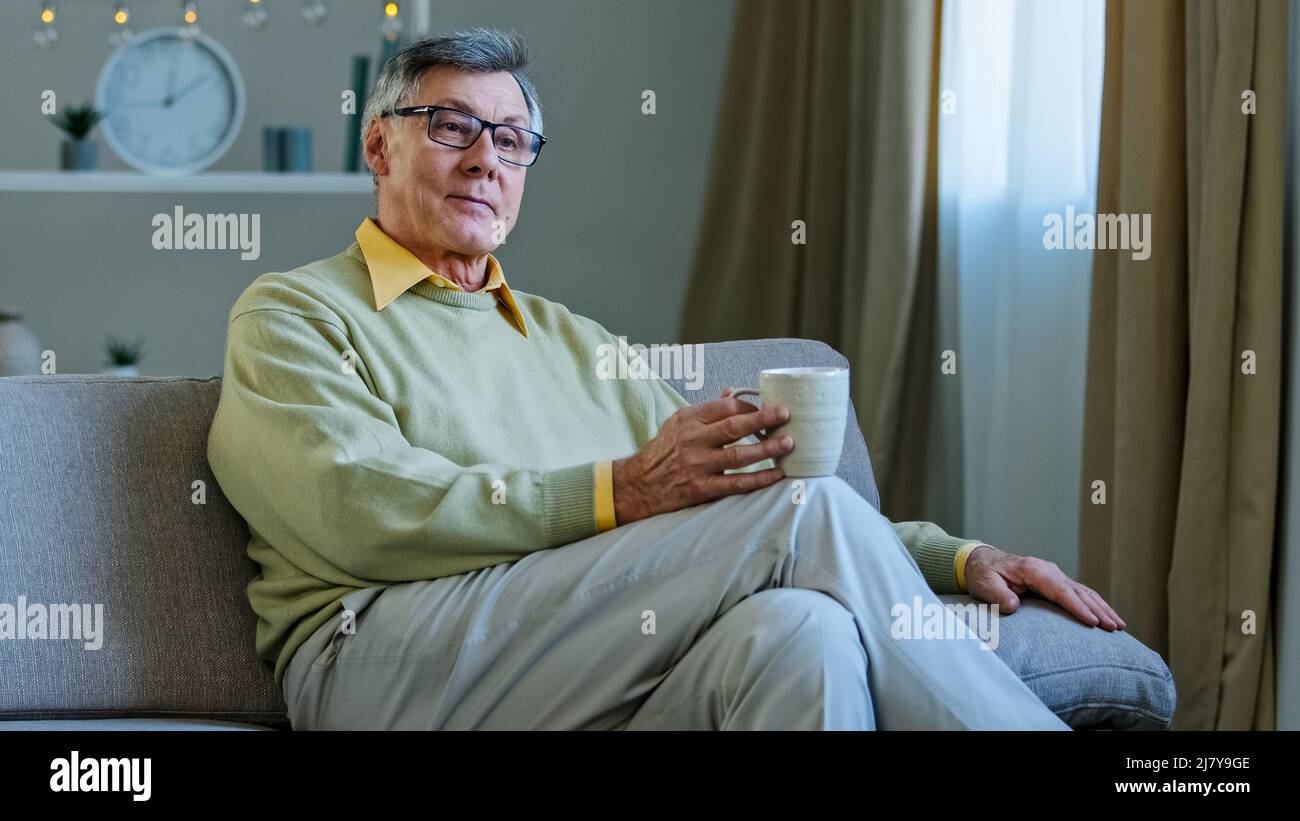 Alter Mann trägt Brille sitzen auf der Couch im Wohnzimmer halten heißen Drink Tasse nachdenklich älteren Opa allein zu Hause entspannen Tee trinken Blick in Stockfoto