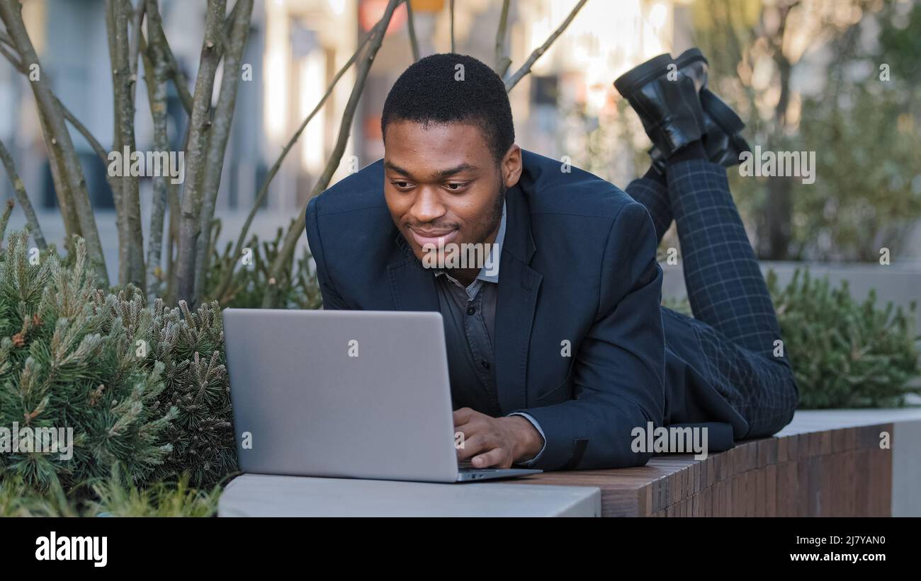 Lächelnd selbstbewusst zufriedener junger Erwachsener Geschäftsmann oder Unternehmensmitarbeiter, der sich hinlegt, nutzt einen Laptop, kommuniziert in sozialen Netzwerken und bereitet Rede vor Stockfoto