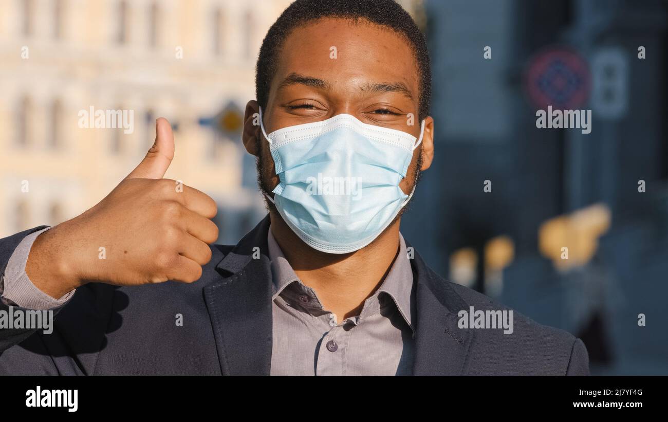 Gemischter Geschäftsmann in Anzug trägt medizinische Gesichtsmaske. Nahaufnahme der Hand, die den Daumen nach oben zeigt. Vorbeugende Vorsichtsmassnahmen aufgrund von Covid-19 Stockfoto