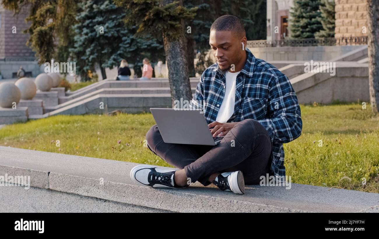 Amerikanischer Student Kerl afrikanischer Geschäftsmann sitzt im Freien in der Stadt Straße auf Laptop-Arbeit online mit Computer E-Learning Musik hören Stockfoto
