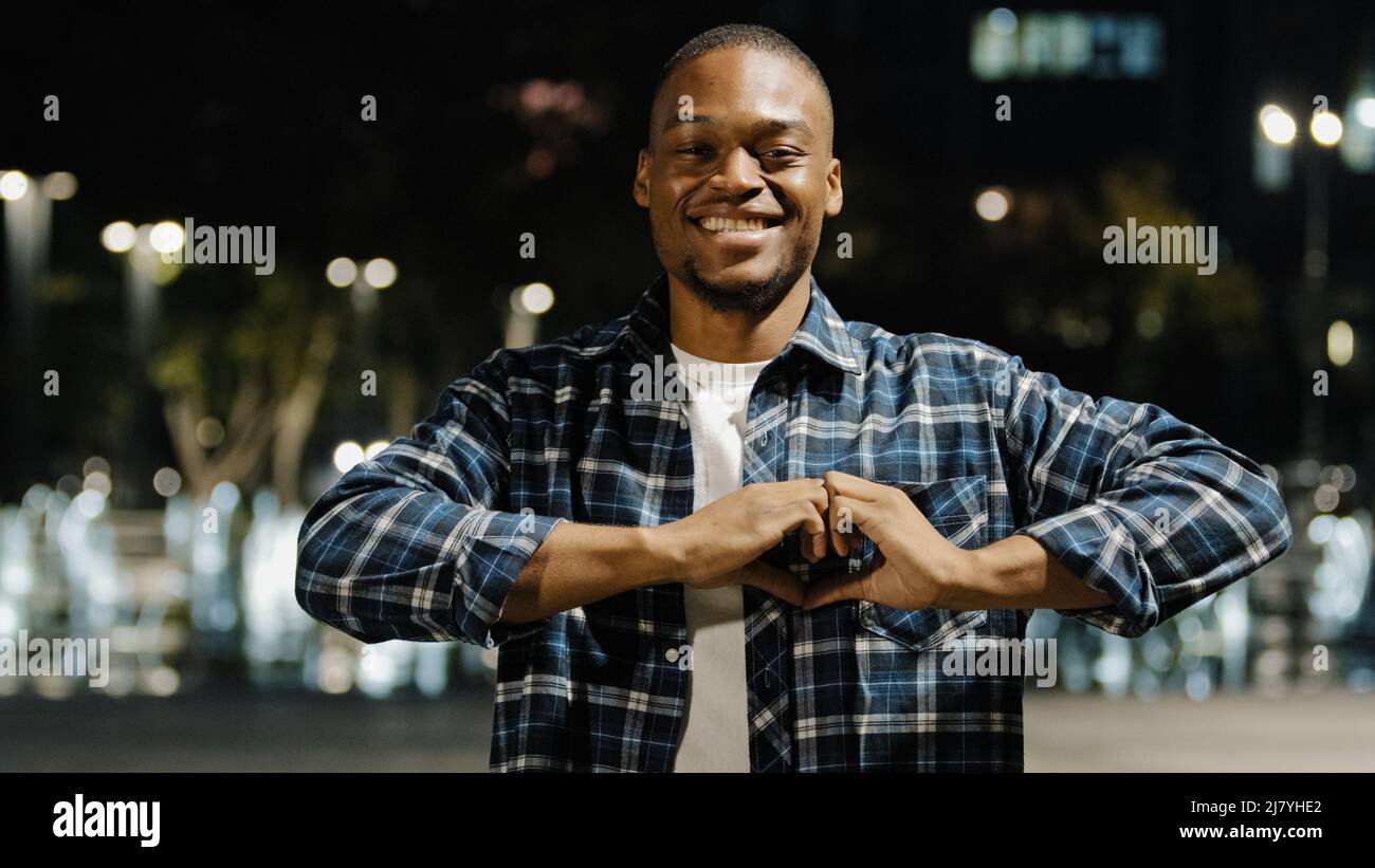 afroamerikanischer Mann Kerl, der in der Nachtstadt steht anhängliches romantisches Porträt, das auf die Kamera schaut, machen Herzform mit den Fingern. Reizender Junge Stockfoto