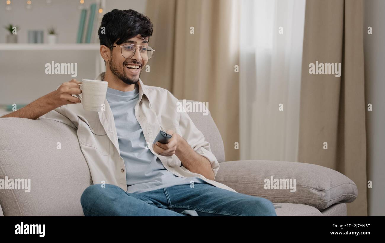 Arabischer Mann bärtiger Kerl mit Brille, die zu Hause auf der Couch sitzt, Tee trinkt, Kaffee schaut lustige Film-Meisterschaft Sport-Spiel-TV jubelt Stockfoto