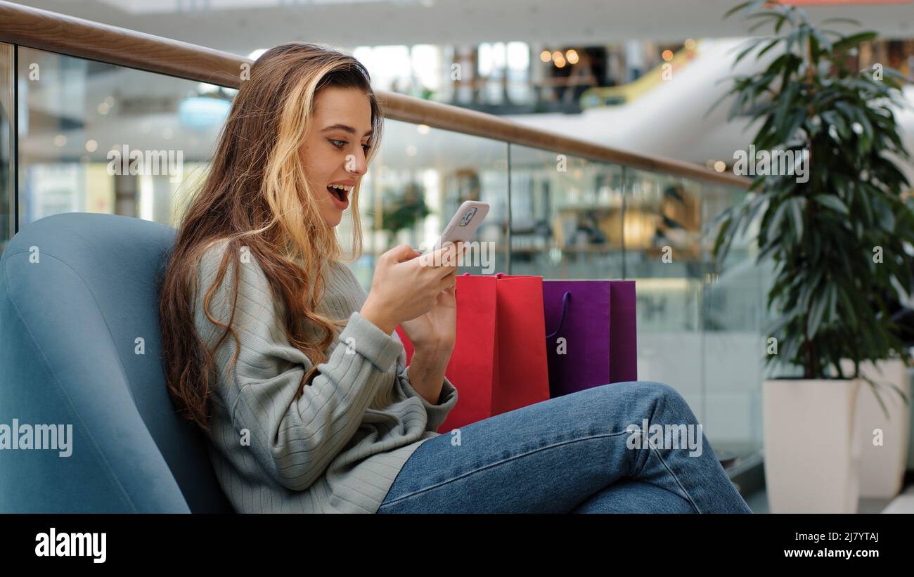 Fröhliche glücklich junge Mädchen Shopper shopaholic Frau sitzt im Einkaufszentrum mit Paketen Blick auf Telefon gewinnt freut Rabatte erfolgreiche Bestellung Stockfoto