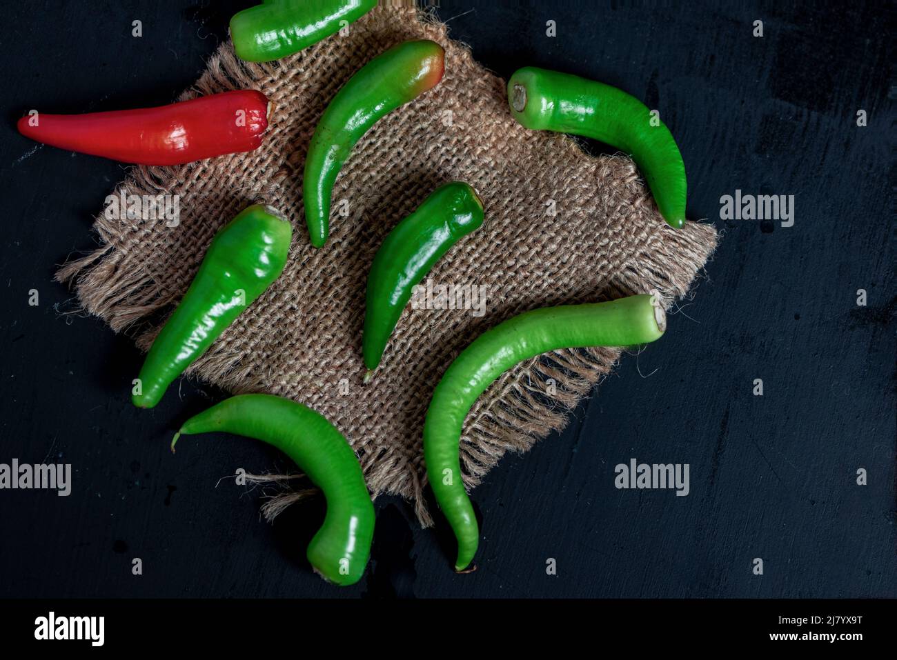 Grüne Chilischoten auf einem hessischen Tischtuch auf schwarzem Holztisch, Nahaufnahme, Stillleben-Fotografie Stockfoto