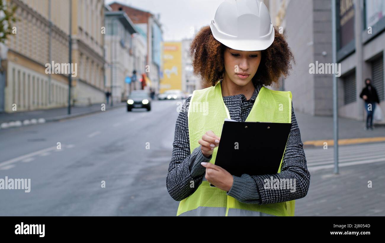 Portrait junge afroamerikanische schöne Ingenieurin mit Sicherheitsweste und Hardhut in der Stadt in der Nähe des Straßenverkehrs, die Daten schreibt Stockfoto