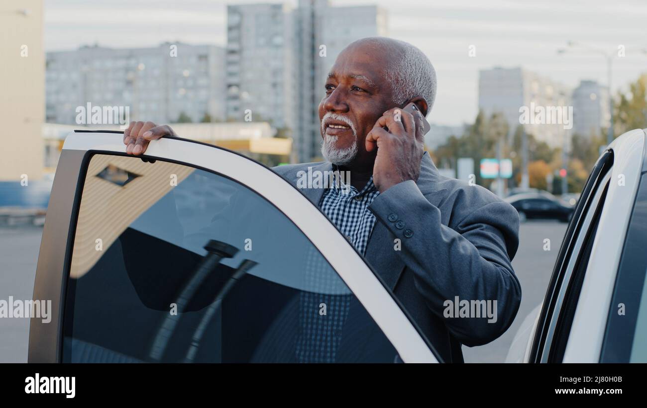 Reifer afroamerikanischer Geschäftsmann, der im Freien in der Nähe des Autos steht und telefonisch telefonische Anrufe entgegennimmt, der Verkaufsvertreter kommuniziert mit dem Kunden auf Stockfoto