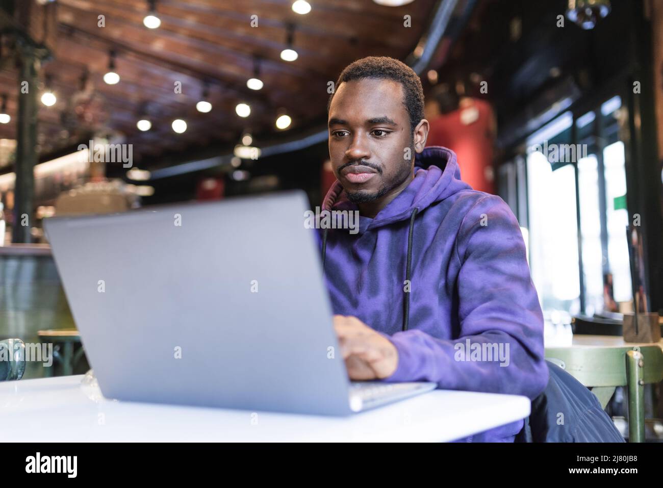 Konzentrierter afroamerikanischer Freiberufler, der einen lässigen Hoodie trägt und am Tisch sitzt und auf Netbook tippt, während er an einem Online-Projekt im Café arbeitet Stockfoto