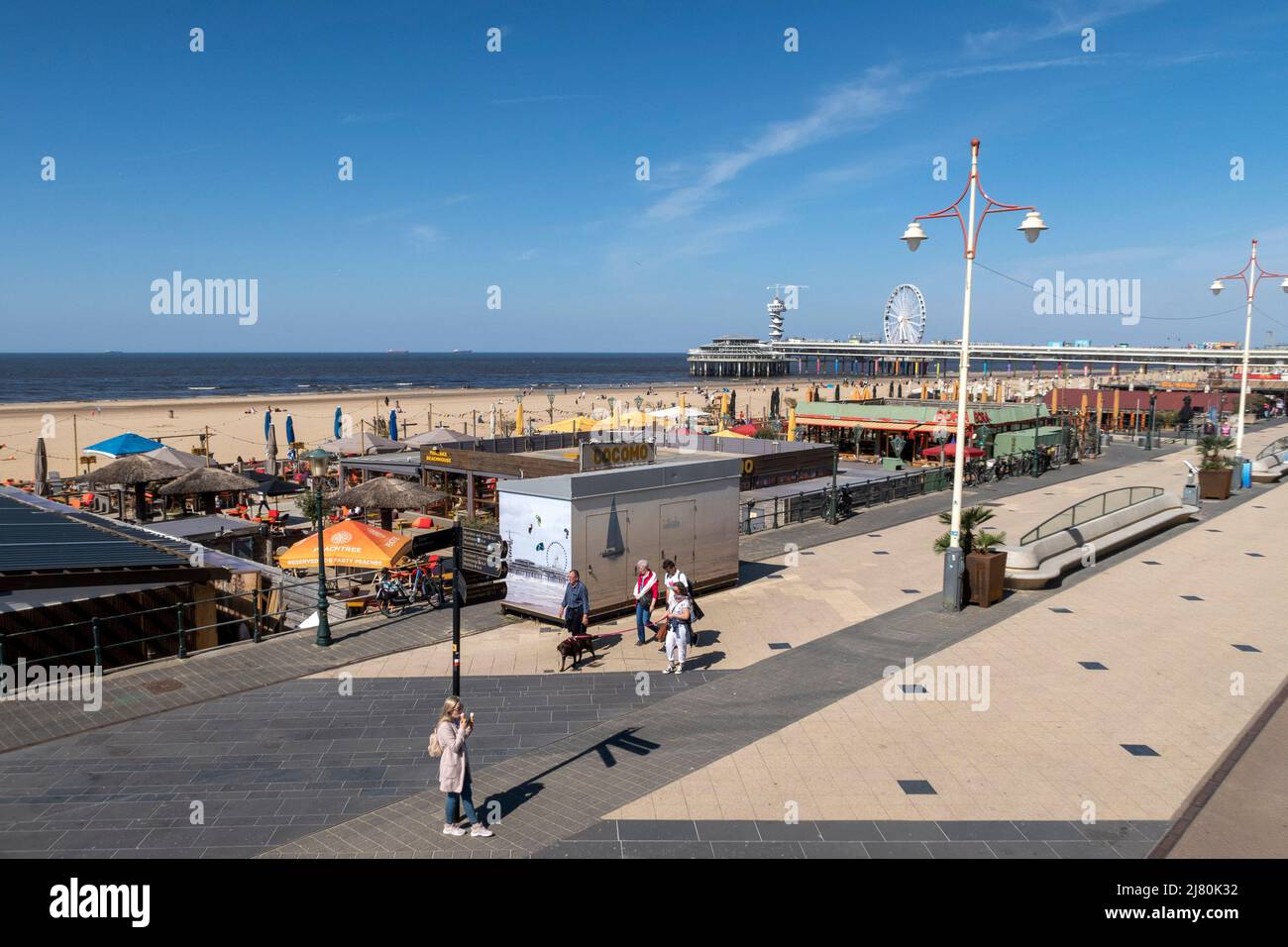 Sonniger Tag am Strand Scheveningen in den Haag, Niederlande, mit Menschen, die entlang der Promenade und dem Pier im Hintergrund spazieren Stockfoto