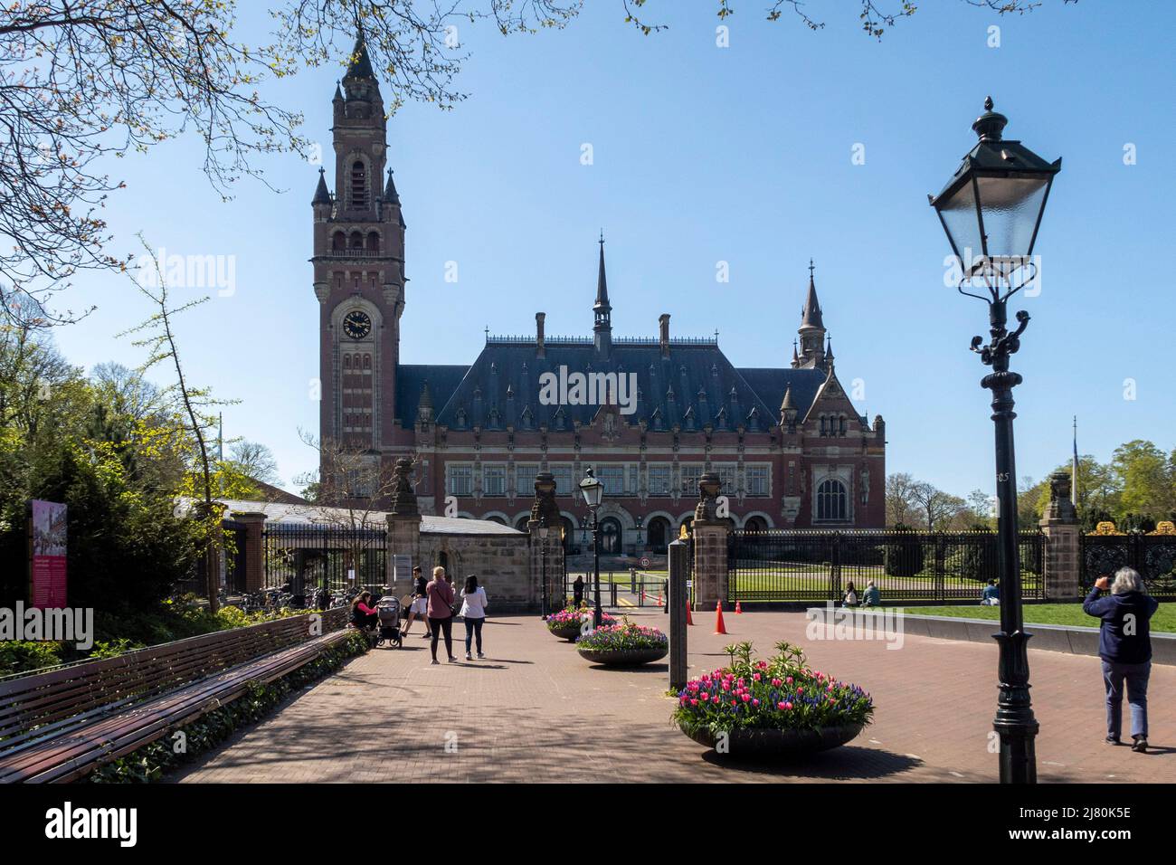 Touristen spazieren und machen Fotos in der Nähe des Friedenspalastes an einem sonnigen Tag in den Haag, Niederlande, umgeben von Frühlingsblumen und Bänken Stockfoto