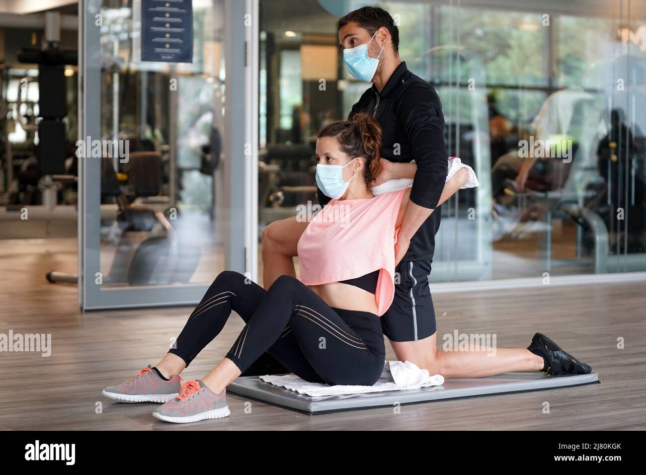 Ein persönlicher Trainer im Fitnessstudio hilft einem Kunden bei der Durchführung von Dehnungsübungen, während er Gesichtsmasken während der Covid-19-Epidemie verwendet Stockfoto