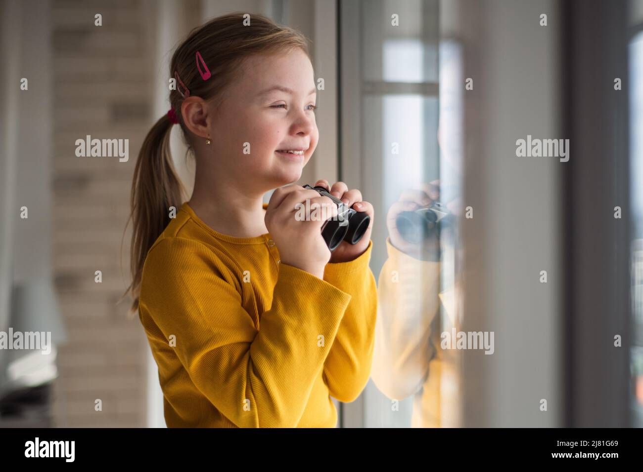 Neugieriges kleines Mädchen mit Down-Syndrom und Fernglas, das zu Hause durch das Fenster schaut. Stockfoto