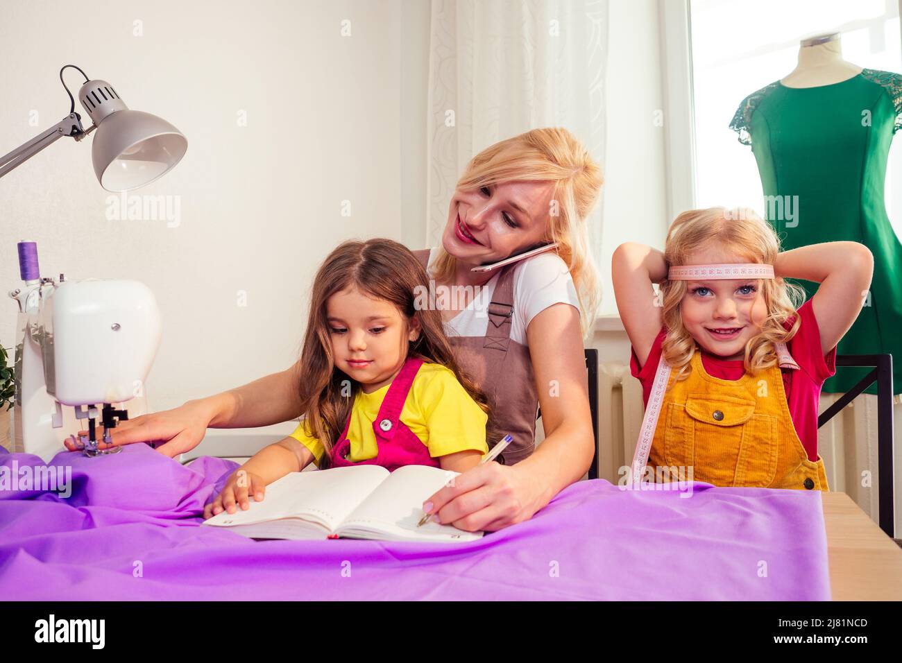 Lächelnde, fröhliche, multitasking-fähige blonde Mutter lehrt eine kleine Schwester, eine Nähmaschine selbst anzunähen und Selfie-Fotos zu machen Stockfoto