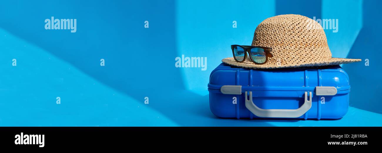 Blauer Plastikkoffer mit Strohhut und Sonnenbrille auf dem Boden in einem hellen Studio für Sommerurlaub Baner platziert Stockfoto