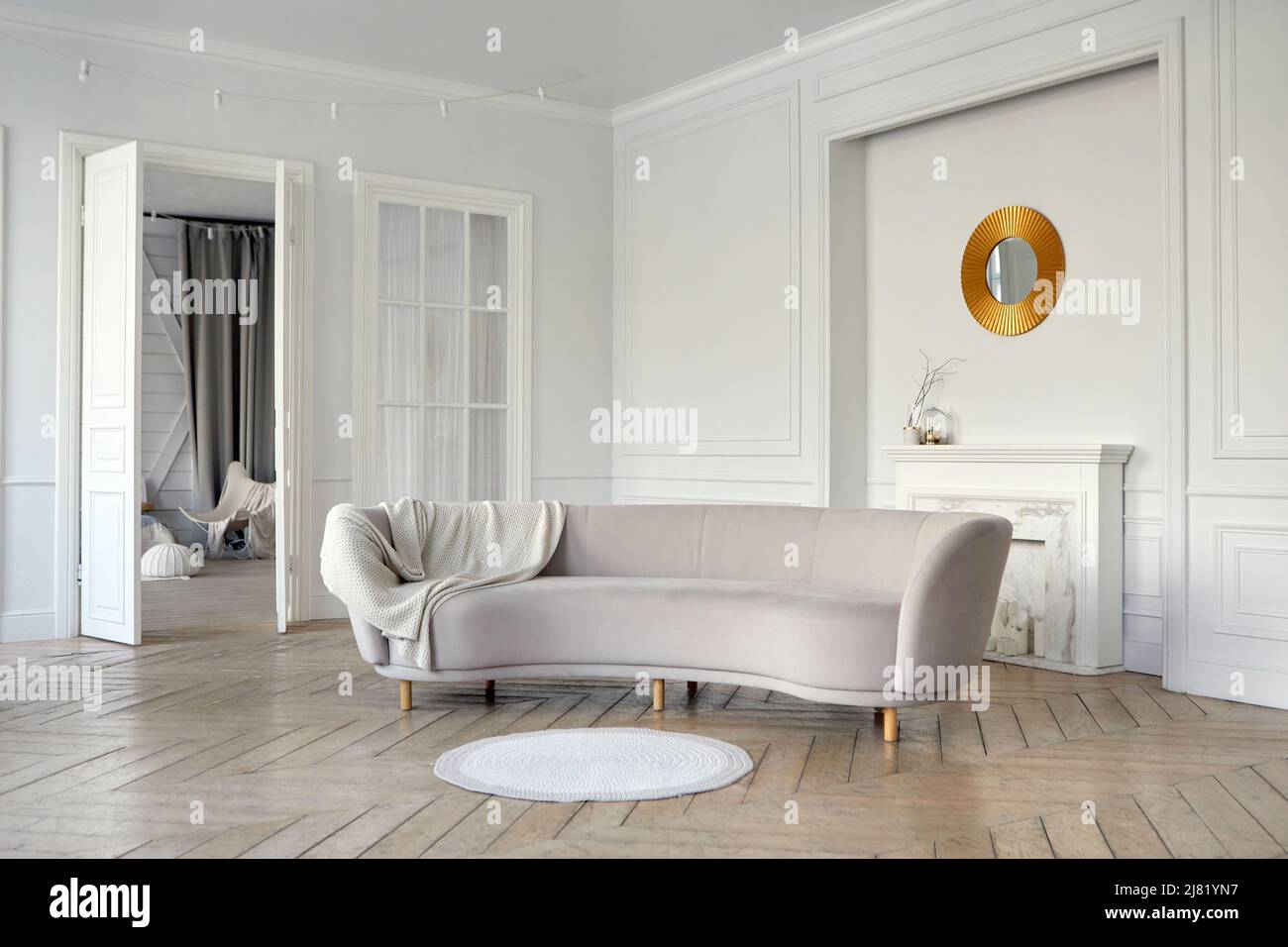 Elegantes Interieur des geräumigen Wohnzimmers mit komfortablem grauem Velours-Sofa und Parkettboden, dekoriert mit künstlichem Kamin und runder Goldeinfassung Stockfoto