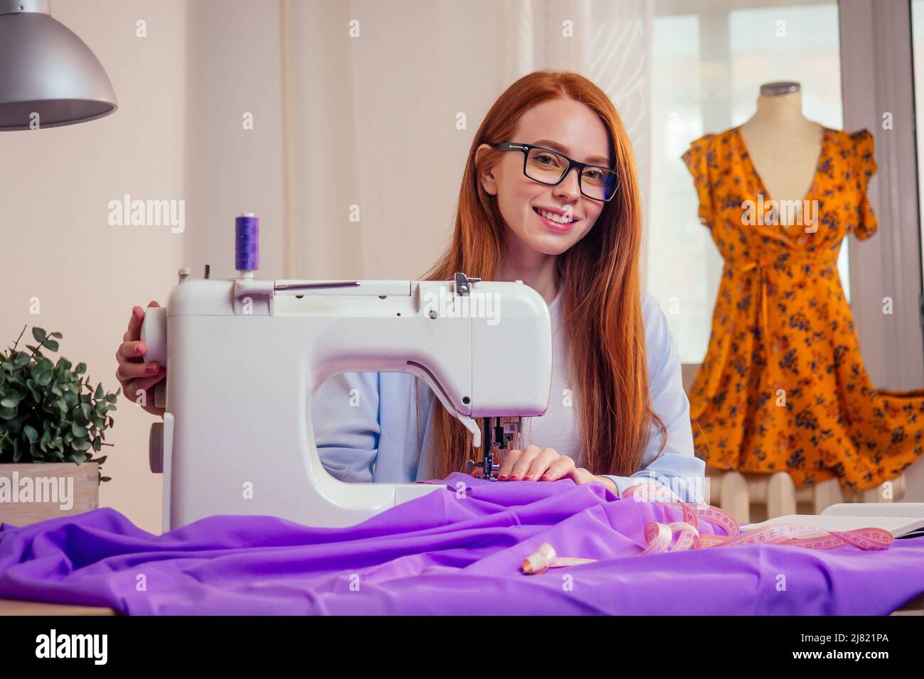 Rothaarige Geschäftsfrau mit Nähmaschine und maniken im Studio-Hintergrund. Schneider schafft eine Kollektion Outfits näht Kleidung in Werkstatt schreiben Stockfoto