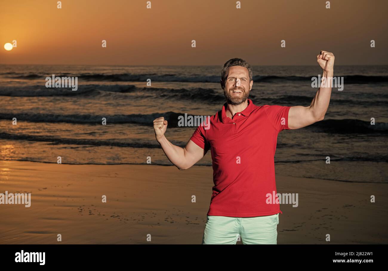 Erfolg feiern. Raum kopieren. Glücklicher Mann mit Bart im Sonnenuntergang über Meer. Erfolgreicher Kerl Stockfoto