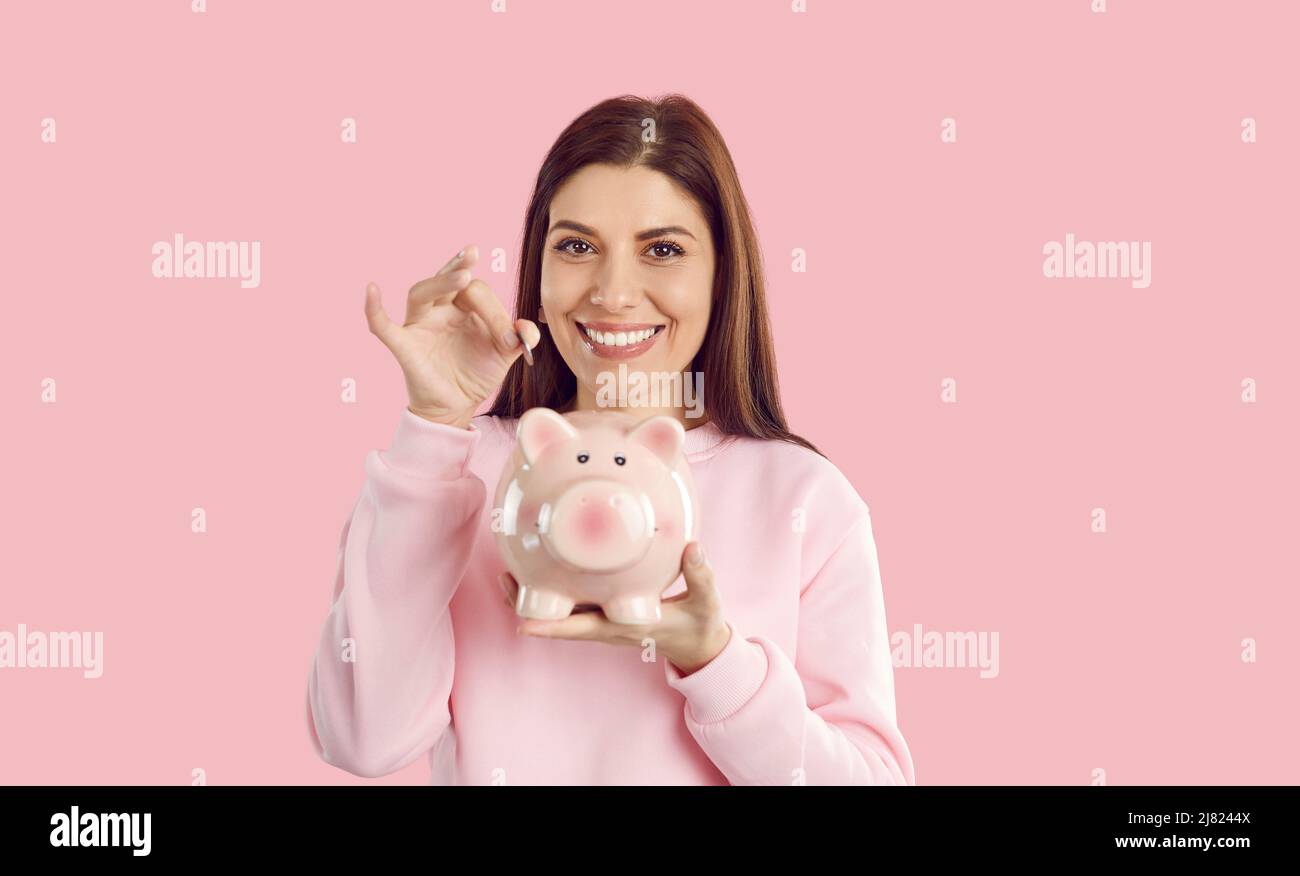 Lächelnde, sparsame Frau wirft eine Münze in ein Sparschwein, das sie auf rosa Hintergrund in der Hand hält. Stockfoto