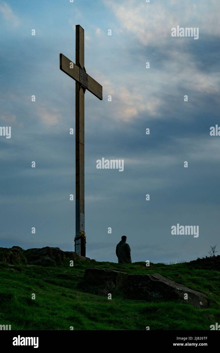 Eine Einzelfigur auf einem Hügel, hoch aufragendes hölzernes Osterkreuz (zur Erinnerung an die Kreuzigung) und Sonnenuntergang am Himmel - The Chevin, Otley, West Yorkshire England Großbritannien. Stockfoto