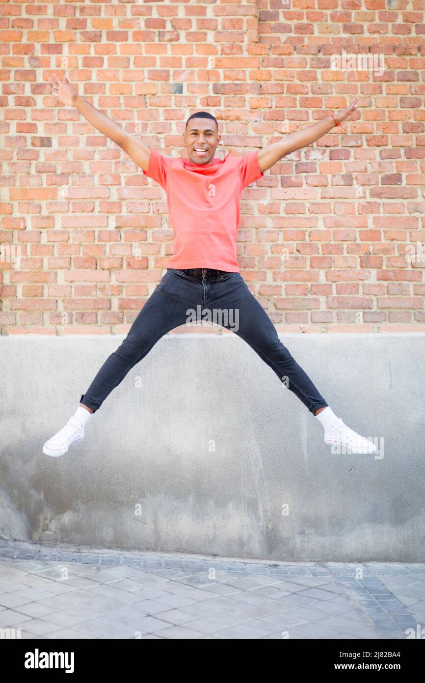 Junger afroamerikanischer Mann, der mit offenen Beinen springt und die Kamera mit einer Wand im Hintergrund anschaut Stockfoto